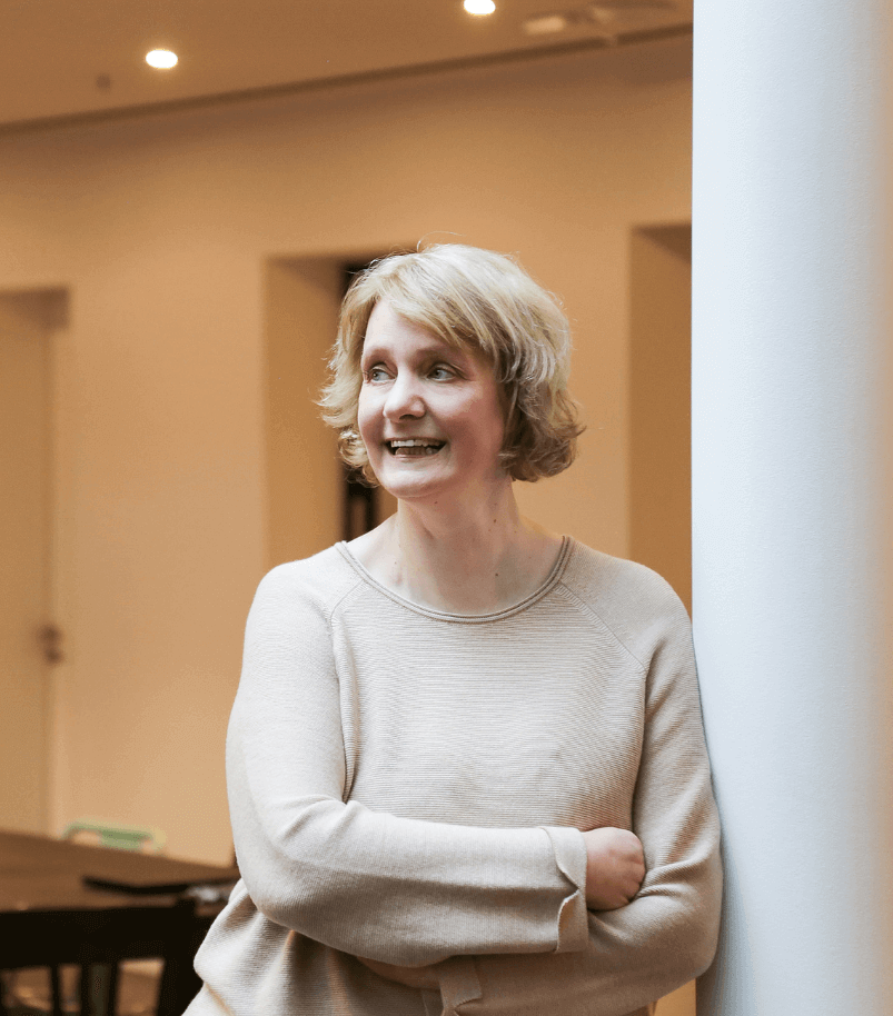 Foto von Peggy Zöllner Düsseldorf. Eine blonde Frau im mittleren Alter steht an eine Säule gelehnt, schaut nach links und lächelt.