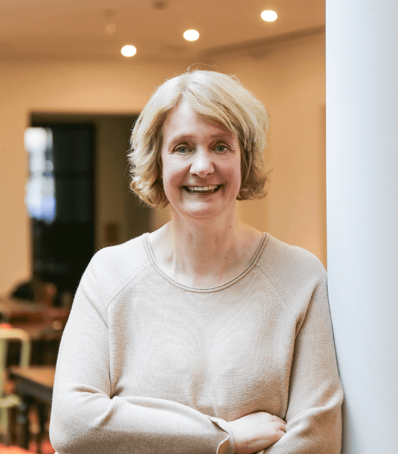 Foto von Peggy Zöllner Düsseldorf. Eine blonde Frau im mittleren Alter steht an eine Säule gelehnt und lächelt.